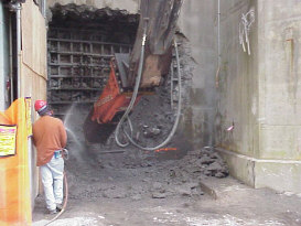 Worker spraying down concrete wall while an excavator jackhammers it